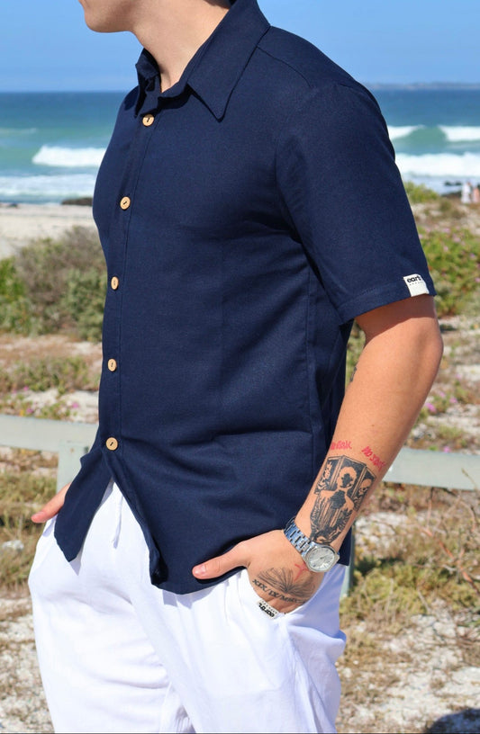 Person wearing a navy linen shirt with white pants standing by the ocean.